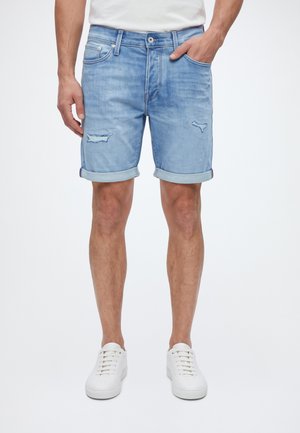 Un homme porte un short en jean bleu clair usé avec des revers roulés, un t-shirt blanc et des baskets blanches sur un fond uni.