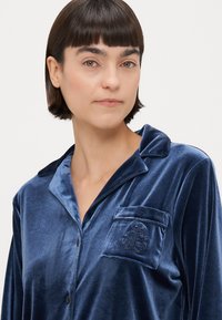 Conjunto de pijama em veludo azul-marinho com um bolso apresentando detalhes bordados, textura suave e design clássico com botões e colarinho.