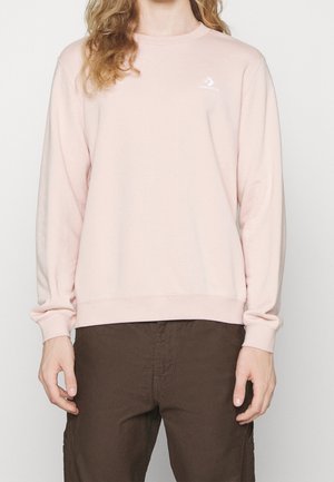 Persoon draagt een lichtroze longsleeve met een klein wit Converse-logo op de borst en donkerbruine broek.