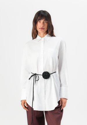 White long-sleeve shirt with a collar, featuring a front pocket, layered over loose brown trousers, accented with a black floral belt.