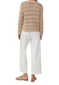 Beige Häkelpullover mit offenem Strickmuster, kombiniert mit weißen weit geschnittenen Hosen und braunen Sandalen mit dekorativen Akzenten.