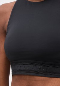 Κοντινό πλάνο ενός ατόμου που φοράει μαύρο αθλητικό μπουστάκι με υψηλό λαιμό και υφασμένη ταινία με την ένδειξη "Ted Baker Sport".