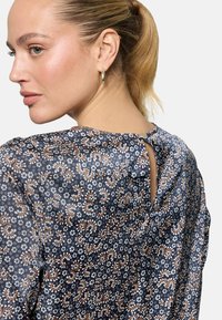 Femme aux cheveux blonds attachés en queue de cheval, portant un chemisier bleu marine à motifs avec une fermeture à boutons dans le dos et des boucles d'oreilles créoles en or.