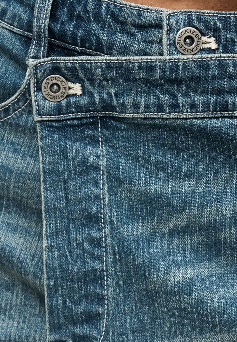 Sezione frontale di jeans in denim blu con cuciture visibili, passanti per cintura e bottoni metallici incisi con il marchio "Dickies".