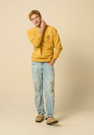 Junger Mann in gelbem Sweatshirt, hellblauen Jeans und beigen Slip-On-Schuhen steht mit einer Hand in der Tasche und der anderen berührt seinen Hals.