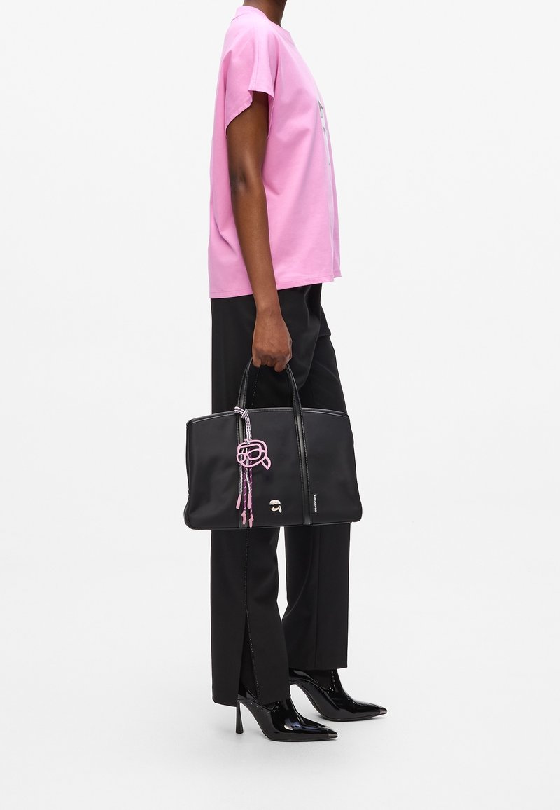 Personne portant un t-shirt rose et un pantalon noir tient un sac à main noir avec un détail en corde rose, chaussée de chaussures noires à talons aiguilles.