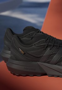 Par sorte atletiksko med teksturerede såler, sorte snørebånd og mærke af Cordura-stof, placeret på en orange overflade.