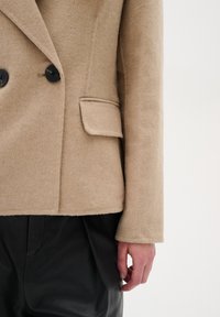 Beige sako z vlněné směsi s černými knoflíky, strukturovanými rameny a jednou náprsní kapsou, v kombinaci s černými koženými kalhotami.