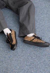 Baskets à motif léopard avec une semelle en caoutchouc noir, design à lacets et accents noirs. Portées avec des chaussettes blanches et un pantalon gris sur un sol bleu.