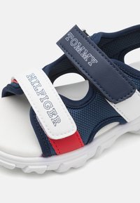 Marinblå sandaler med vita och röda detaljer, texturerade remmar och justerbara kardborrestängningar. Lätta med en dämpad sula.