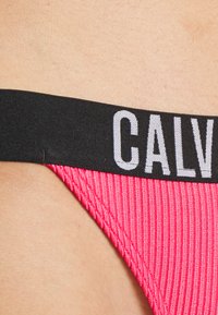 Růžové žebrované tanga s černým elastickým pasem, na kterém je vytištěno "CALVIN KLEIN" bílou barvou. Hladká textura, minimalistický design.