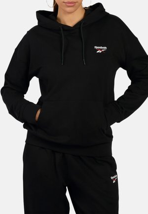 Frau trägt schwarzen Reebok-Hoodie und passende schwarze Jogginghose mit Logo auf Brust und Oberschenkel, Hände in den Taschen des Hoodies.