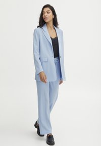 Svetlo modra jakna z izrezom in sprednjimi žepi, v kombinaciji z ustreznimi hlačami. Pod njo je črna čipkasta majica in črni loaferji.