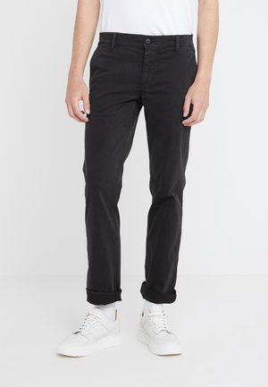 Man met zwarte omslagbroek, witte sokken en witte sneakers staand tegen een effen lichte achtergrond.