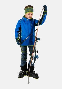 Veste de ski bleue avec des accents noirs et verts, associée à un pantalon de ski bleu marine. L'enfant tient un bâton de ski et porte des chaussures de ski noires. Bandeau texturé.