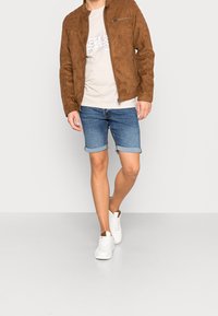 Brun mockakjorta, beige grafisk t-shirt, denimshorts med upprullade fållar och vita sneakers. Outfiten har en avslappnad, lager-på-lager look.