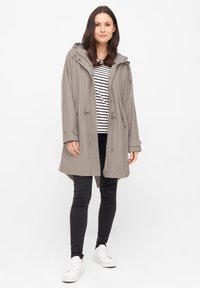 Grijze lichte regenjas met een capuchon, stropdas in de taille en drukkers, gedragen over een gestreepte lange mouwtop en zwarte broek.