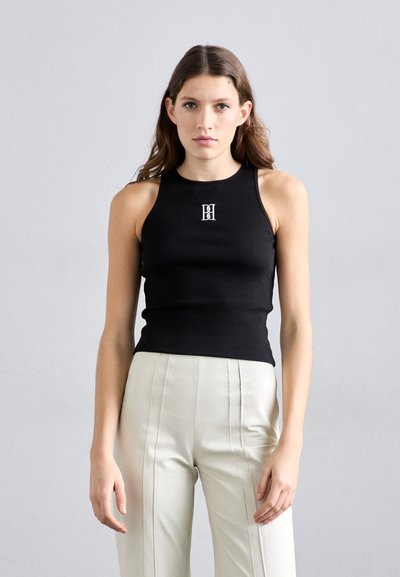 Schwarzes, geripptes Tanktop mit hohem Kragen, das ein kleines weißes Logo in der Mitte trägt. Kombiniert mit hellen, hochgeschnittenen Hosen.