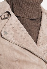 Béžová semišová bunda s perforovaným klopou a stříbrnými kovovými doplňky, vrstvená přes hnědý žebrovaný rolák. Jsou viditelné textilní textury.