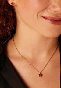Gouden ketting met een kleine bloemenhanger met rode stenen, gecombineerd met bijpassende oorbellen met rode stenen, gedragen op blote huid.