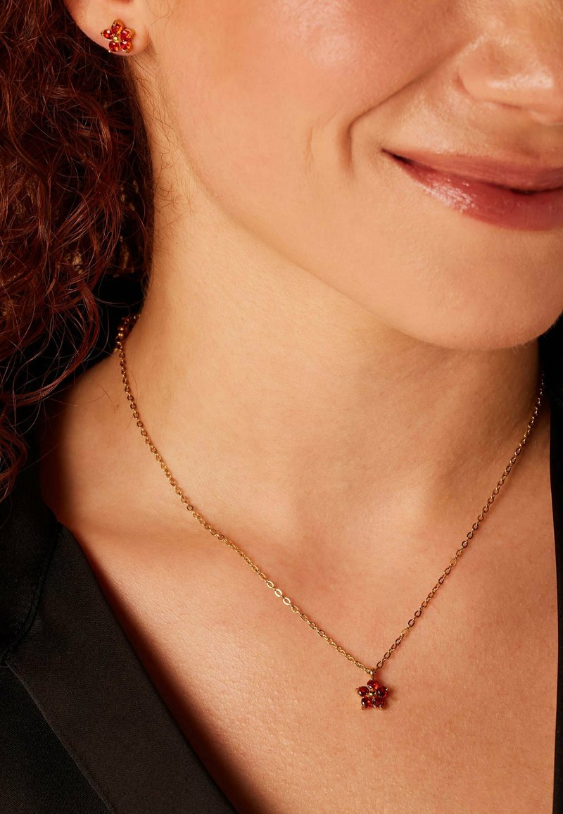 Gouden ketting met een kleine bloemenhanger met rode stenen, gecombineerd met bijpassende oorbellen met rode stenen, gedragen op blote huid.