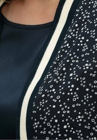 Cardigan navy con accenti bianchi, caratterizzato da una sezione decorata con piccole forme e stelle. Texture liscia e design classico.
