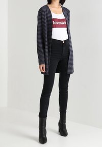 Gilet rayé bleu marine sur un débardeur blanc avec le texte "lovesick", assorti à un jean noir taille haute et des bottines noires.