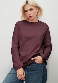 Hosszú ujjú bordó pulóver kerek nyakkal és laza szabással, kék farmernadrággal párosítva. Puha tapintású, minimális varrási részletekkel.