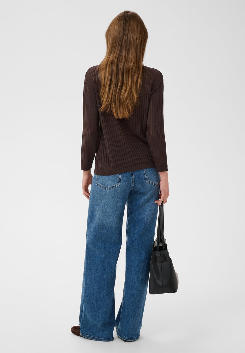 Femme aux longs cheveux bruns portant un pull marron foncé et un jean bleu à jambes larges, tenant un sac à main noir, debout de dos.
