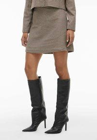 Mini suknja smeđe boje s uzorkom ptičje kosti i uskim krojem, uparena s crnim kožnim čizmama na visoku petu do koljena.