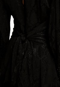Robe noire avec tissu texturé légèrement brillant, taille cintrée par un nœud noué, et manches longues et fluides.