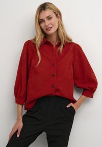 Chemise en velours côtelé rouge avec un devant à boutons, des manches bouffantes et une coupe décontractée, associée à un pantalon noir à taille élastique.