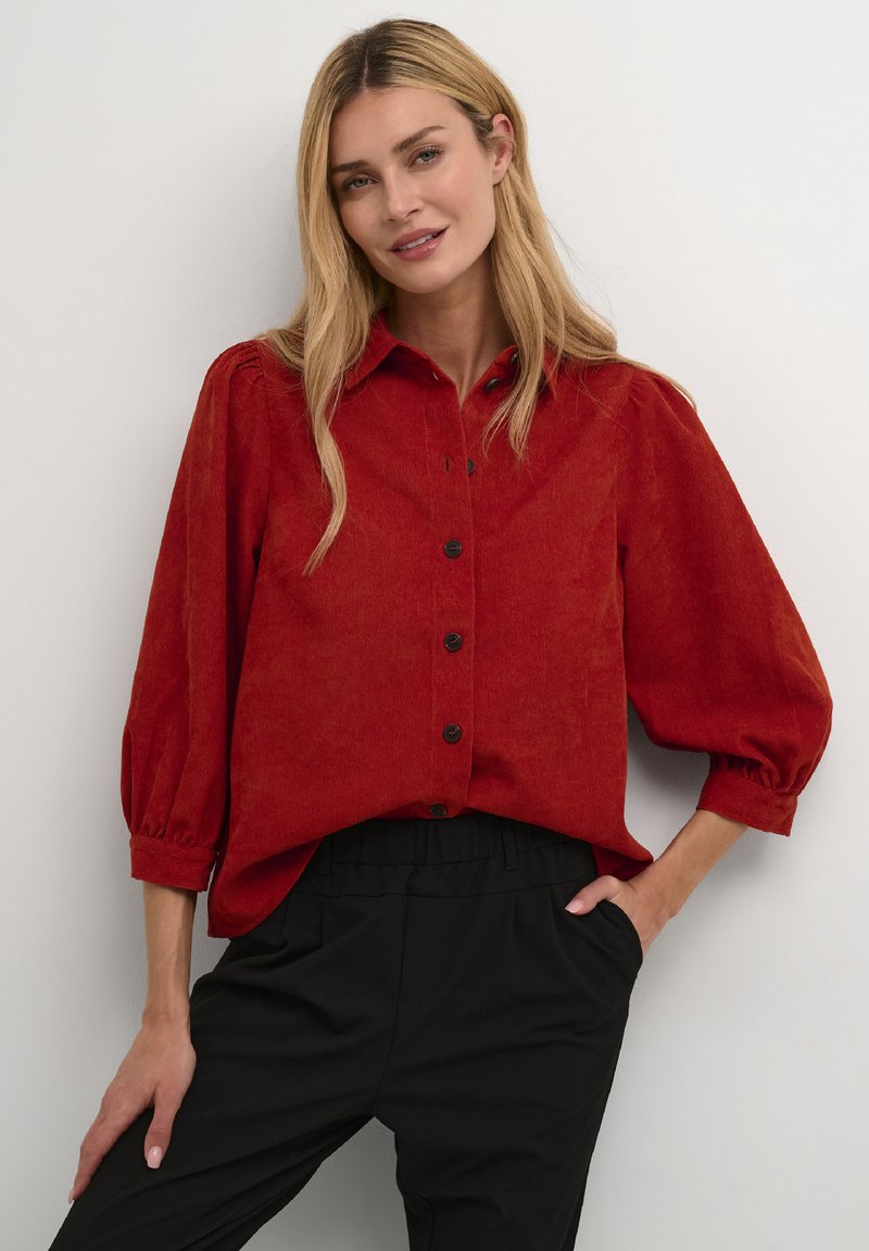 Chemise en velours côtelé rouge avec un devant à boutons, des manches bouffantes et une coupe décontractée, associée à un pantalon noir à taille élastique.