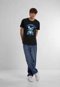 Melnā T-krekls ar zila lāča grafiku un uzrakstu "Favorita" priekšpusē, apvienots ar ziliem džinsiem un baltām kedām. Vienkāršs dizains.