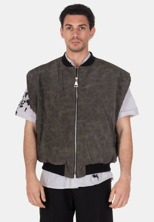 Uomo che indossa un gilet verde scuro oversize senza maniche con zip sopra una camicia a righe a maniche corte, in piedi davanti a uno sfondo semplice.