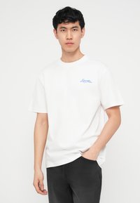 Νεαρός άνδρας με λευκό μπλουζάκι με μικρό μπλε κείμενο στο στήθος και μαύρο παντελόνι, στέκεται μπροστά σε απλό ανοιχτόχρωμο φόντο με το ένα χέρι στην τσέπη.