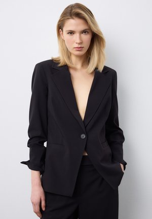 Blonde Frau, die einen schwarzen, taillierten Blazer mit einem Knopf trägt, die Ärmel leicht hochgekrempelt, und passende schwarze Hosen, stehend vor einer weißen Wand.