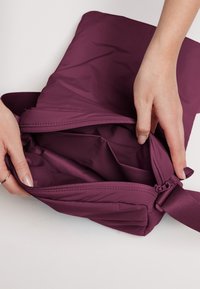 Borsa di nylon viola con chiusura a zip, caratterizzata da una texture liscia e una tracolla. L'interno mostra un ampio spazio e nessuna tasca aggiuntiva.