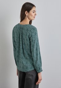 Grön mönstrad blus med ett geometriskt mönster, långa ärmar och rund halsringning. Tyget har en slät struktur och en lätt lyster.