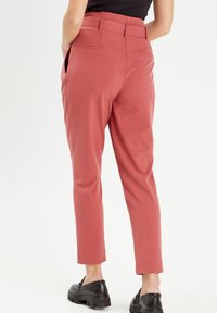 Pantalon tailleur rouge avec une texture lisse, jambes fuselées et poches latérales, associé à des chaussures à lacets noires.