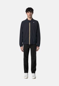 Giacca bomber blu con zip frontale, polsini e orlo elastici; presenta una striscia gialla e bianca al centro. Indossata con pantaloni neri.