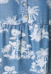 Denimstoff mit einem hellblauen Hintergrund und weißen tropischen Palmenbaumprints. Enthält Knopfdetails und einen gerafften Saum.