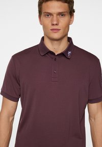 Burgundy pikétröja i mjuk tygkvalitet, med krage, treknappars placket och en liten blå detalj på kragen.