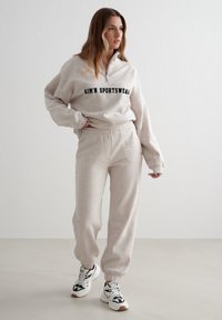 Sudadera beige claro con cuello con cremallera y logo negro "AIM'N SPORTSWEAR", combinada con pantalones jogger a juego y zapatillas blancas con detalles en negro.