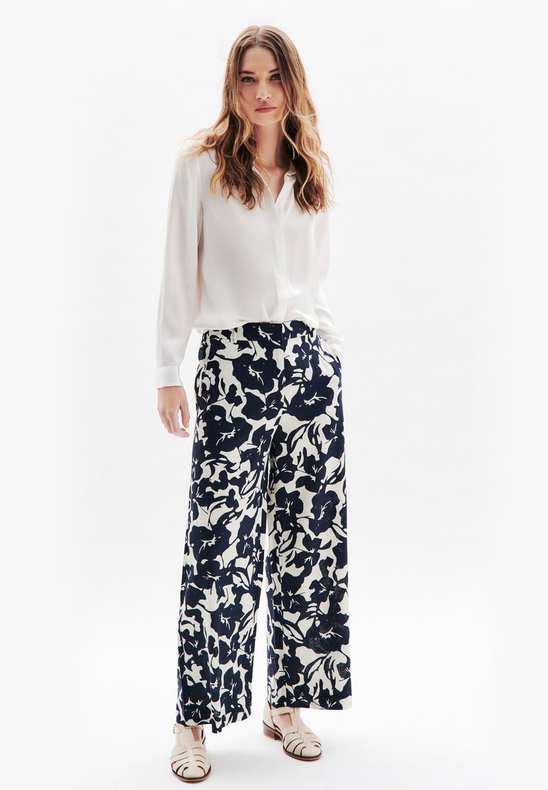 Vit långärmad blus i kombination med vida marinblå byxor med blommönster. Modellen bär ljusa platta sandaler.