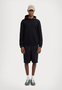 Svart hettegenser med frontlomme og snor, kombinert med matchende shorts. Beigefarget caps og grønne sneakers med hvite detaljer fullfører stilen.