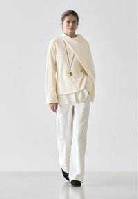 Cream oversized sweater with a draped design, layered over a white shirt, paired with white wide-leg pants and black shoes.