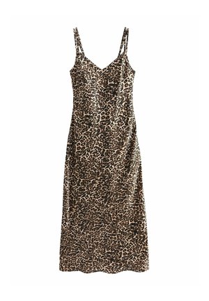 Mujer con vestido midi sin mangas con estampado de leopardo y botas blancas hasta el tobillo, de pie contra una pared beige con las manos detrás de la espalda.