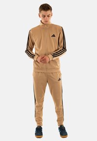 Giovane in piedi, che indossa una tuta Adidas beige con strisce nere su maniche e pantaloni, e sneakers blu scuro con suole color gomma.