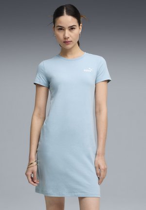 Kobieta o ciemnych włosach w niebieskiej sukience Puma z krótkim rękawem stojąca na tle szarego gradientu.